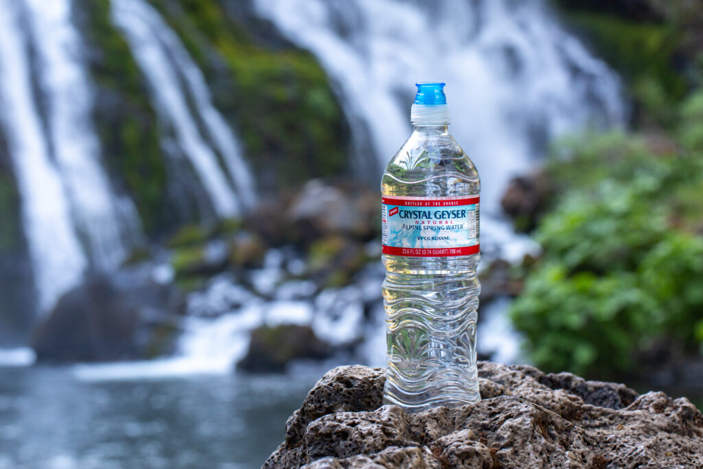 A Crystal Geyser water bottle on a rock with greenery and a waterfall in the background.