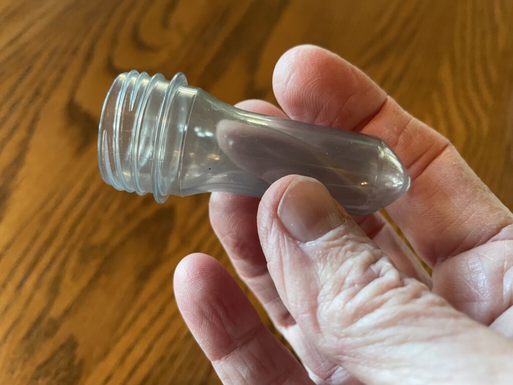A hand holding a plastic bottle preform, which is a tube with one closed end, and the other end has the screw-threads of the top of a recycled plastic bottle